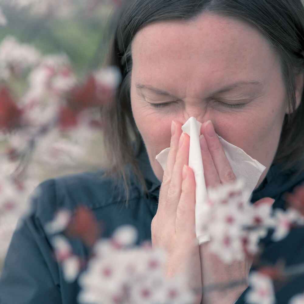 Woman sneezing into a tissue due to allergies and being surrounded by flowers